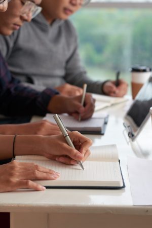 Close-up shot of Asian university students writing test while sitting at desk of modern classroom, blurred background